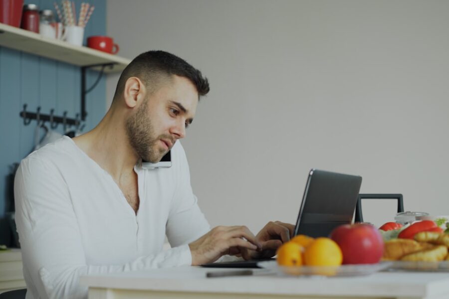 Man on phone and laptop at kitchen table.